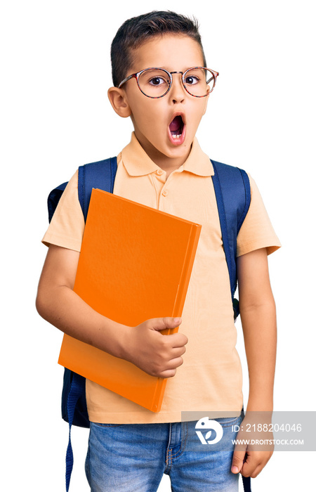 Little cute boy kid wearing school bag and holding book scared and amazed with open mouth for surprise, disbelief face