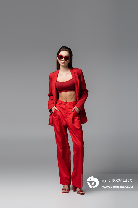 full length of young woman in trendy red suit and sunglasses standing with hands in pockets on dark grey.