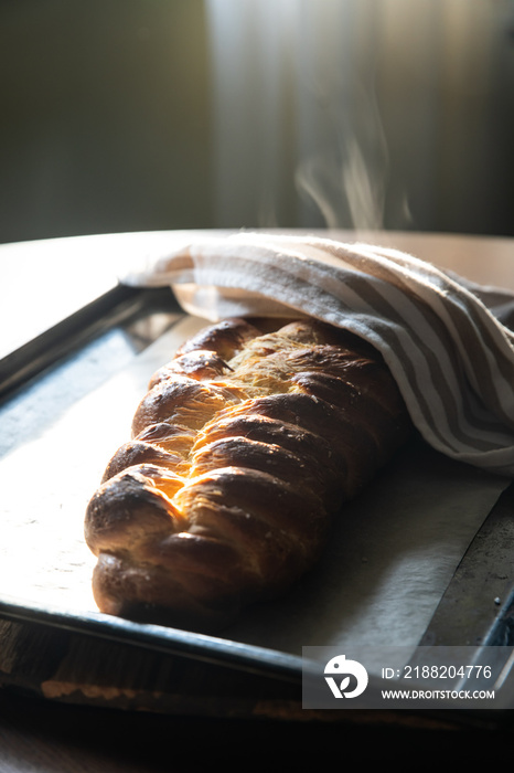 Steaming homemade braided bread (challah) half covered with a towel just from oven on wooden board