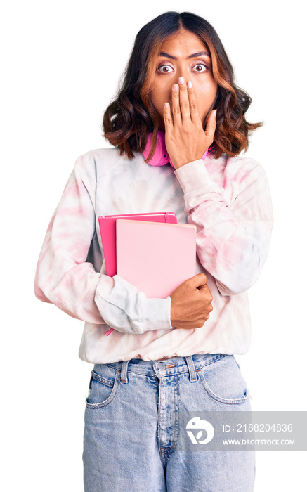 Young beautiful mixed race woman holding books wearing headphones covering mouth with hand, shocked and afraid for mistake. surprised expression