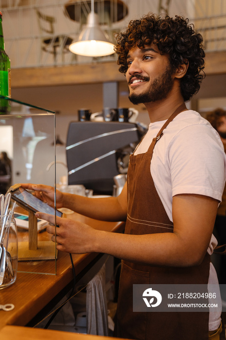 Young indian smiling handsome curly barista working with tablet