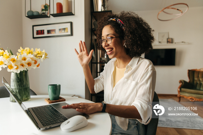 Friendly young african girl smiles and waves at laptop camera sitting at table. Brunette with afro hair wears white shirt. Concept of use