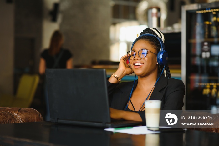 Happy black girl in wireless headphones studying online, using laptop and taking notes, cafe interior, copy space