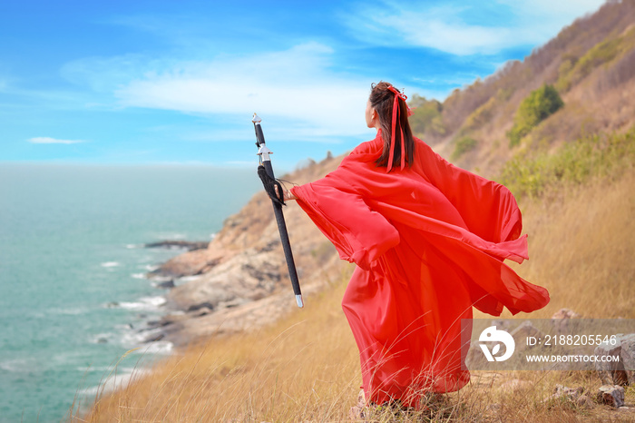 Rear view portrait of young and beautiful woman wearing red Chinese warrior costume with black sword, she post using sword on mountain with sea and nature outdoor