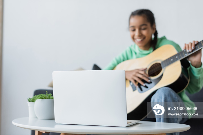 blurred african american teenage girl smiling while learning to play acoustic guitar near laptop