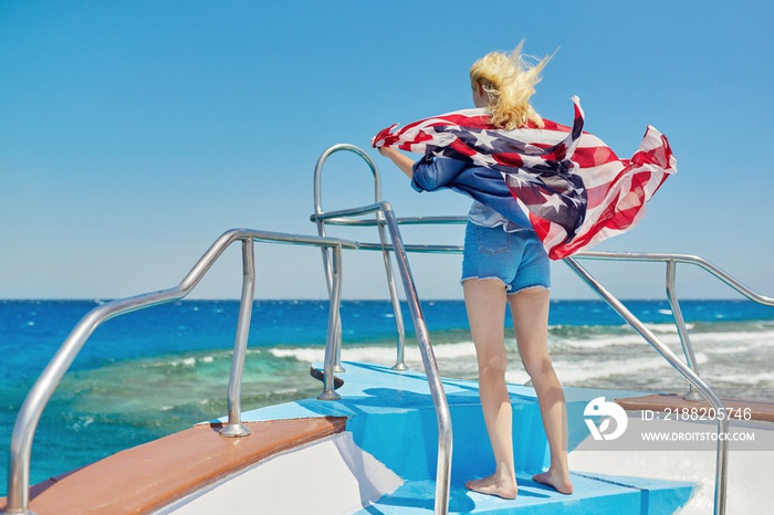 Young blonde woman in denim with flag of America on shoulders, back view, in sea