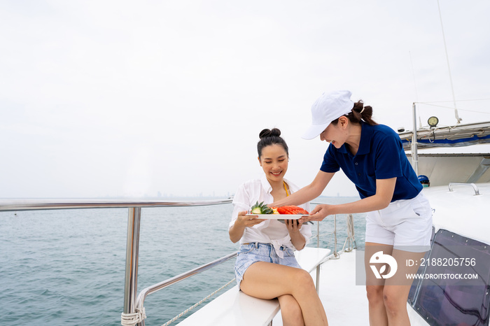 Asian woman waitress serving fresh fruit to passenger tourist on luxury private catamaran boat yacht sailing in the ocean. Attractive girl enjoy outdoor lifestyle travel on summer holiday vacation.