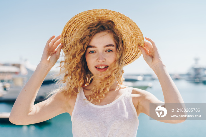 lovely girl with doll beauty holds a hat on her head with two hands on the background of ships and the ocean