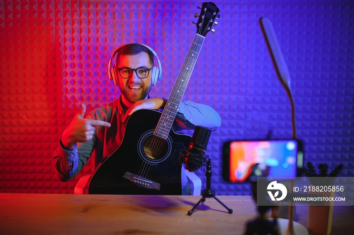 A young stylish guy with a beard wearing casual or music teacher playing guitar in front of smartphone camera. Online guitar training or vocal lesson