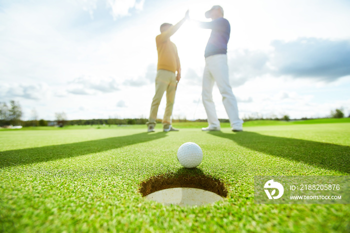 Golf ball lying on green play field close to hole with two successful players making high five on background