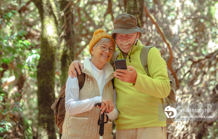 Lovely caucasian senior couple with backpack hiking together in the woods enjoying healthy lifestyle and retirement while using mobile phone