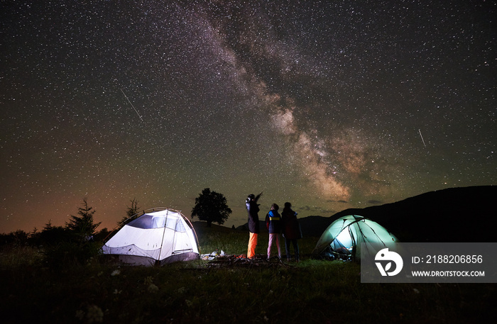 Rear view mother and two children hikers resting at night camping in mountains, standing beside campfire and two glowing tents, enjoying incredible starry sky and Milky way. Woman pointing at sky