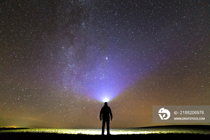 Black silhouette of man with head flashlight on grassy field under beautiful dark summer starry sky.