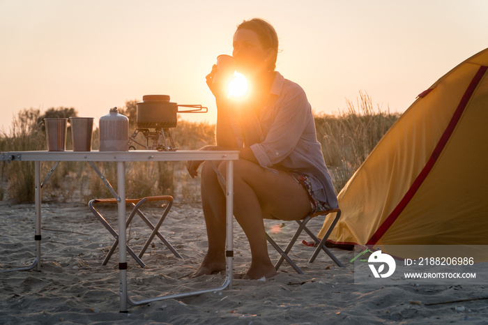 Sunset scenic view of woman holding metal glass. Foldable camping gas fire system with a  pot on a table.