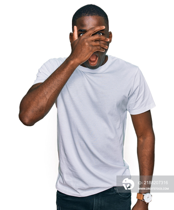 Young african american man wearing casual white t shirt peeking in shock covering face and eyes with hand, looking through fingers with embarrassed expression.