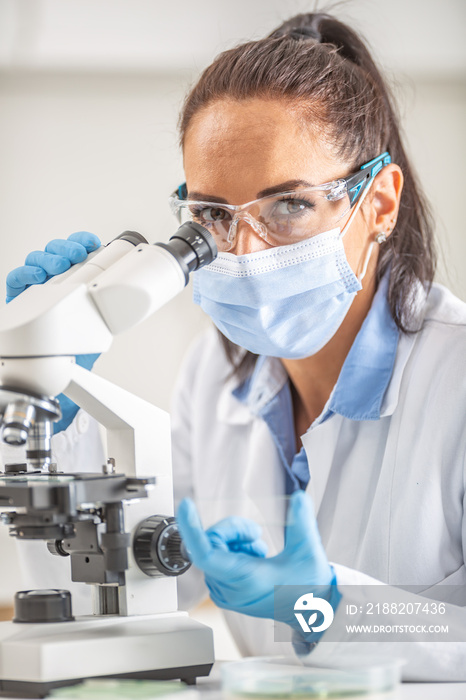 Female lab technician in protective glasses, gloves and face mask sits next to a microscope and conical flask, looking into the camera