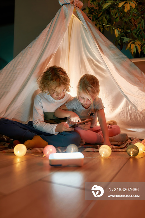 Two adorable little kids, brother and sister using tablet pc while sitting on a blanket in a teepee made with bedsheets at home