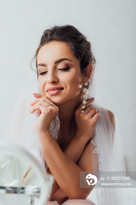 Selective focus of young bride with closed eyes touching lace veil near mirror