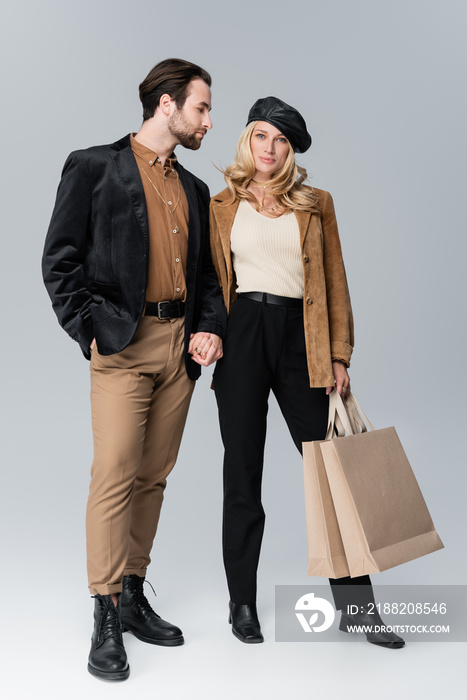 bearded man holding hands with blonde woman in stylish beret standing with shopping bags on grey