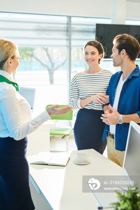 Content friendly bank representative in formalwear standing at desk with computer and open diary and gesturing hands while welcoming young couple in bank office