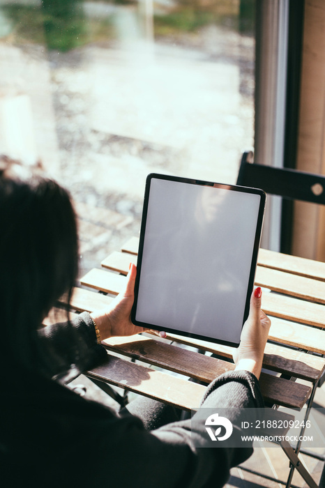 Woman with red nails holding an iPad in a bright room on a wooden table near the window. Work from home