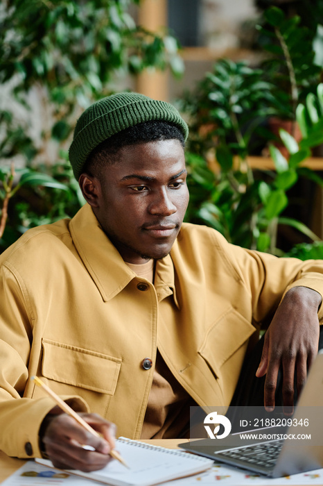 Young serious African American manager making notes on paper while sitting by workplace in front of laptop and planning work