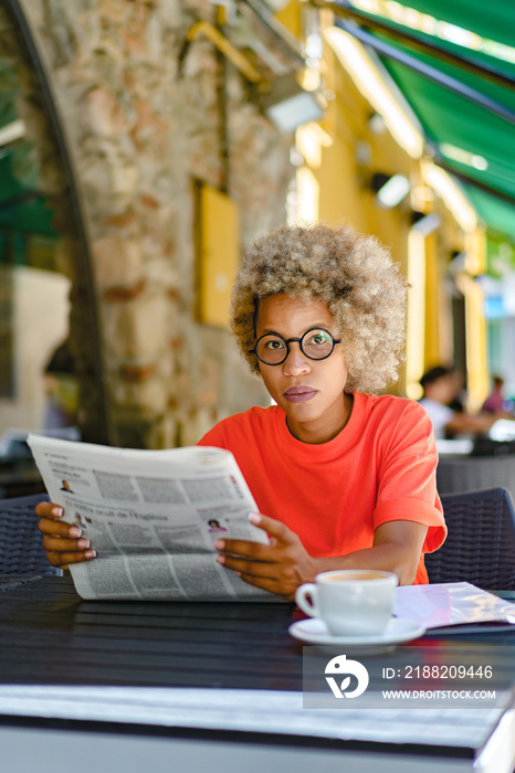 Woman with an afro hairstyle drinking coffee and reading newspaper at cafe. Leisure and news concept