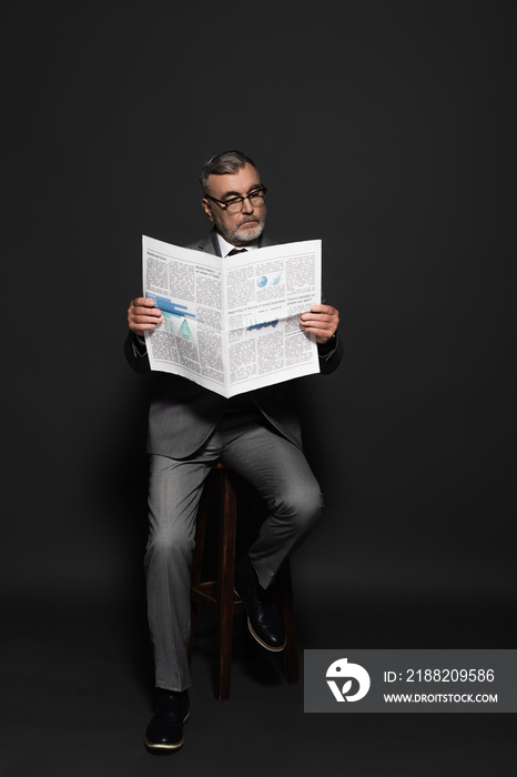 full length view of man in suit and eyeglasses sitting on high stool and reading newspaper on dark grey.