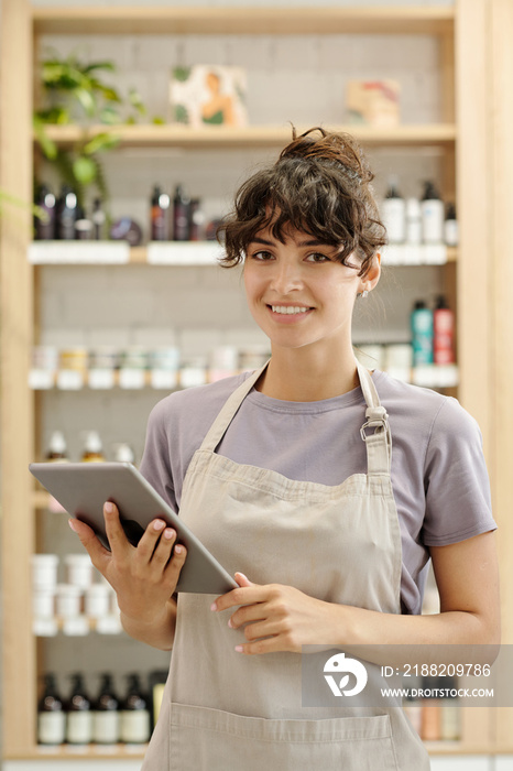 Young successful female shop assistant with digital tablet standing in front of camera against displays with beautycare products