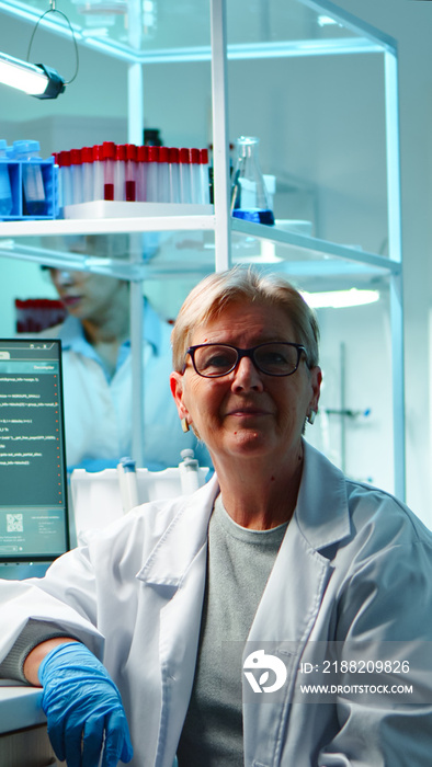 Portrait of senior scientist woman looking exhausted at camera sitting in equipped lab late at night. Team examining virus evolution using high tech for scientific research, vaccine development