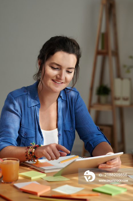 Pretty smiling woman reading a book on table computer and taking notes