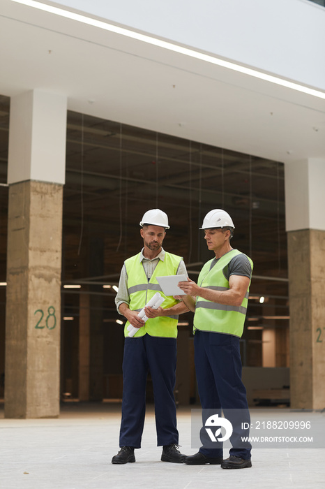 Vertical full length portrait of two building contractors using digital tablet while standing at construction site, copy space above