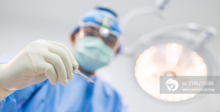 Close up of surgeon’s hand holding a scalpel and blurred male doctor’s face on background with copy space. Selective focus at blade knife inside modern operating room in hospital.Medical concept.