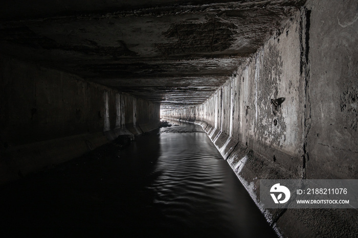 A square concrete drainage tunnel with a light at the end that shines from the turn of the tunnel.