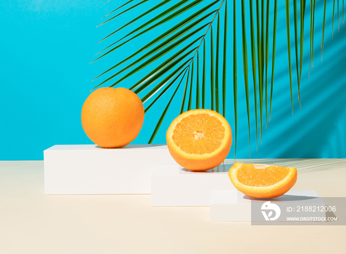 Modern still life with fresh and ripe orange fruit on podium or pedestal on a blue background with tropical palm leaf. Minimal detox diet or summer concept.