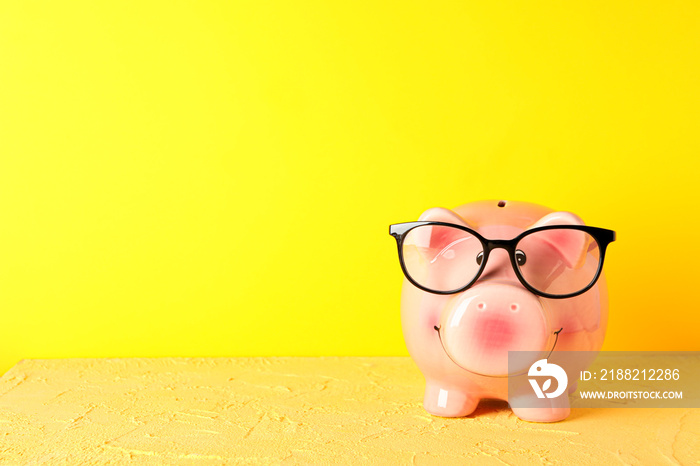 Happy piggy bank with glasses on color table against yellow background, space for text. Finance, saving money