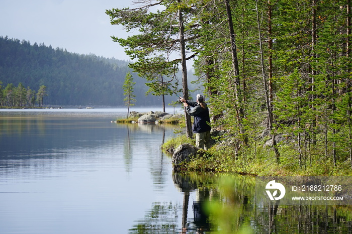 Man casting a fishing rod, at the lake, trees around it, reflections on the water. Summer in Lapland, Finland