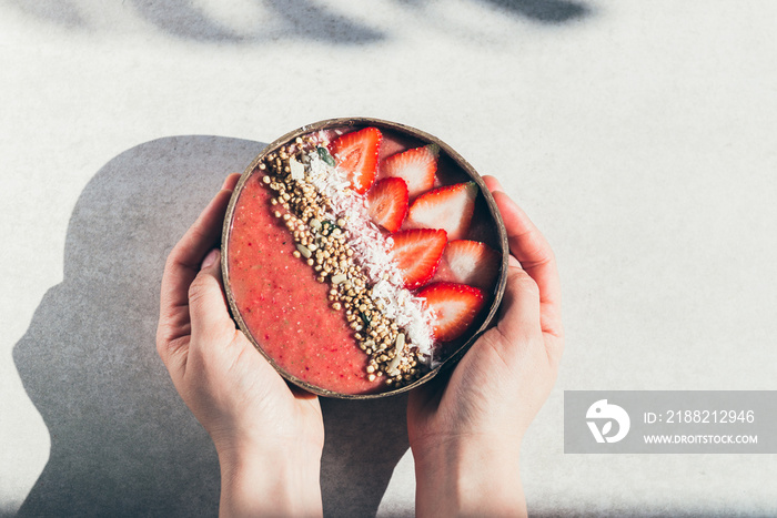 Pink Smoothie from Banana and Strawberries with pieces of Strawberries, Granola and Coconut Shreds on top in Coconut Shell Bowl, strong shadows. Healthy summer breakfast, top view