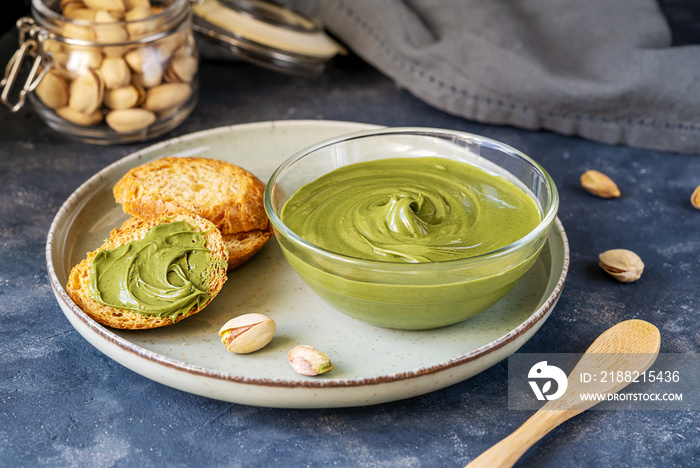 Pistachio cream in glass bowl with crostini on plate, napkin, wooden tea spoon, raw pistachios in jar on dark stone background