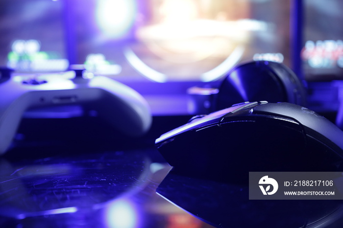 A gaming desk setup with screens illuminating a gamepad and mouse and a gaming laptop on the background with lit keyboard keys