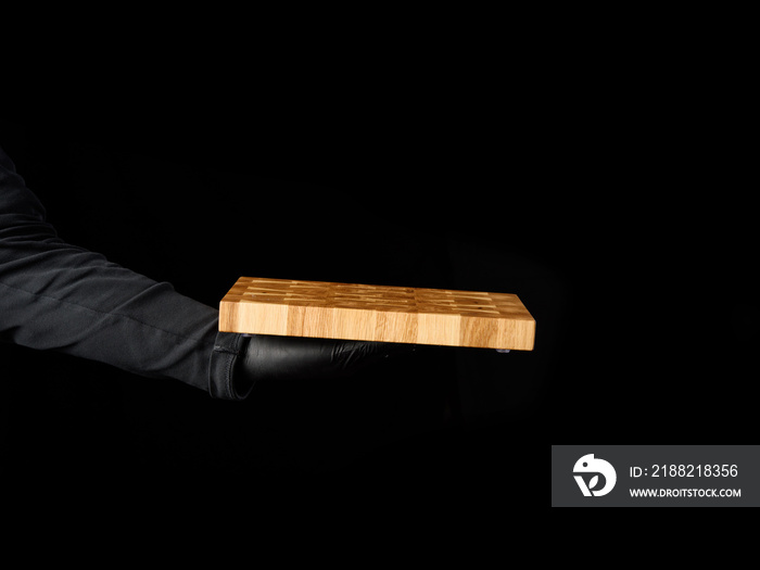 male chef black clothes stands against a black background and holds in his hand a empty rectangular wooden kitchen chopping board