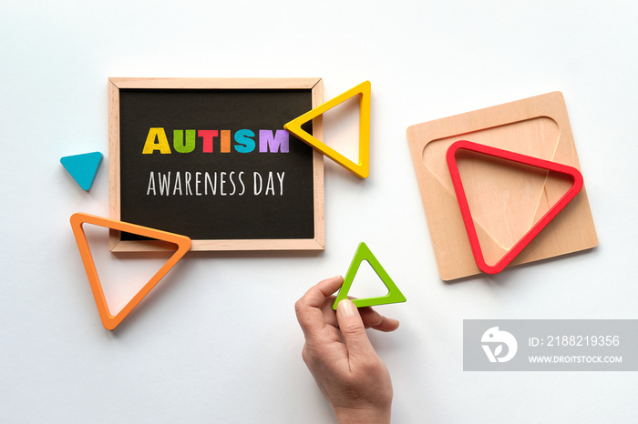 Text Autism awareness month on blackboard. Nested wood triangles in rainbow colors, human hand. Geometric education stacking puzzle. Montessori sensory toy. Flat lay on white paper background.