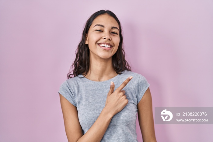 Young brazilian woman wearing casual t shirt over pink background cheerful with a smile of face pointing with hand and finger up to the side with happy and natural expression on face