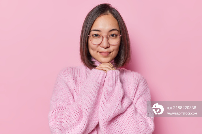 Portrait of charming beautiful Asian woman keeps hands under chin has healthy skin looks directly at camera wears transparent eyeglasses and warm knitted jumper isolated over pink background