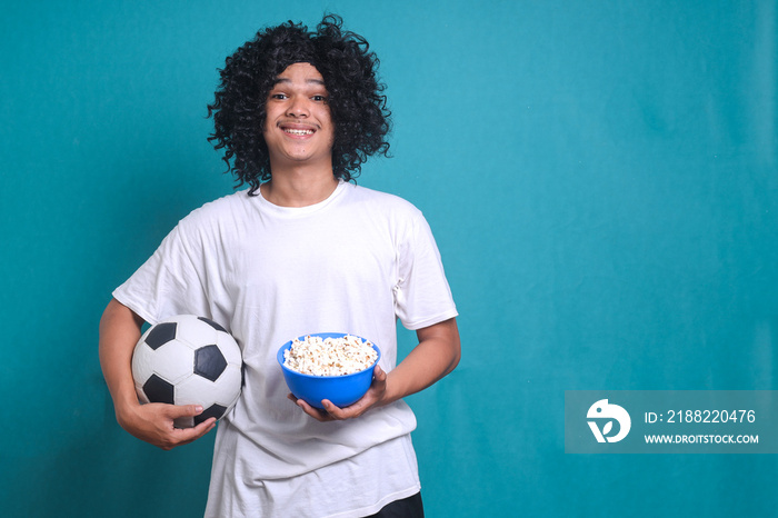 Happy football fan watching tv game while holding popcorn bucket and hold soccer ball