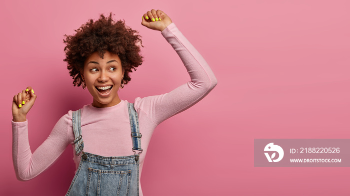 Portrait of optimistic dark skinned young woman poses with hands up, dances carefree, wears turtleneck and overalls, looks aside, isolated over pastel rosy wall, copy space for your promotion