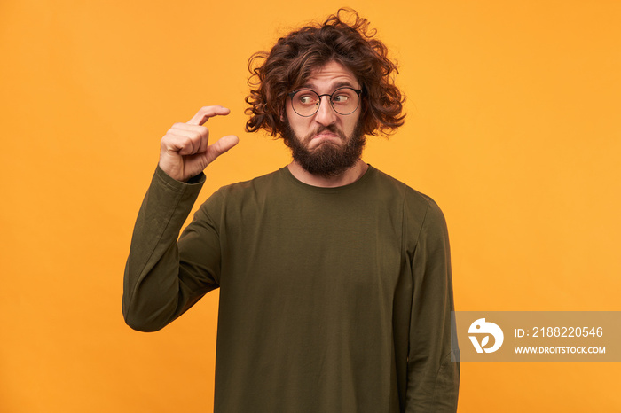 Indoor photo of guy demonstrating small thing.Handsome bearded male with dark curly hair, shows something tiny with hands, looking upset with indignation on it, isolated over yellow background.