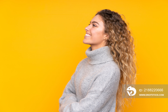 Young blonde woman with curly hair wearing a turtleneck sweater isolated on yellow background in lateral position