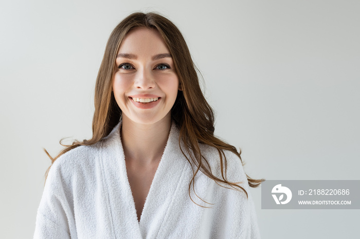 cheerful woman in white bathrobe looking at camera and smiling isolated on grey