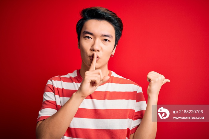 Young handsome chinese man wearing casual striped t-shirt standing over red background asking to be quiet with finger on lips pointing with hand to the side. Silence and secret concept.